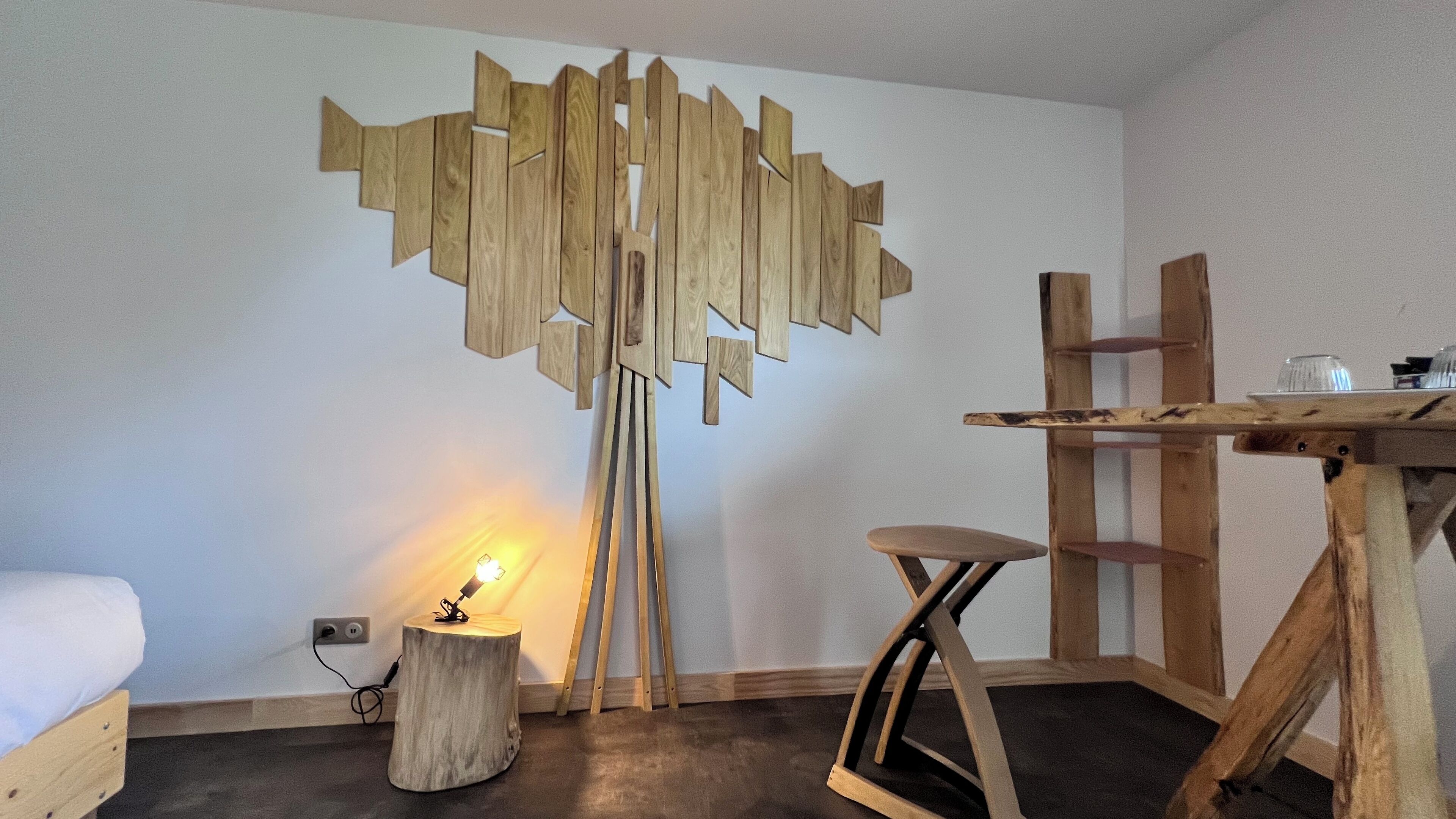 A room with a bed, a wooden table with shelves, a stool, and a unique wooden wall art piece.