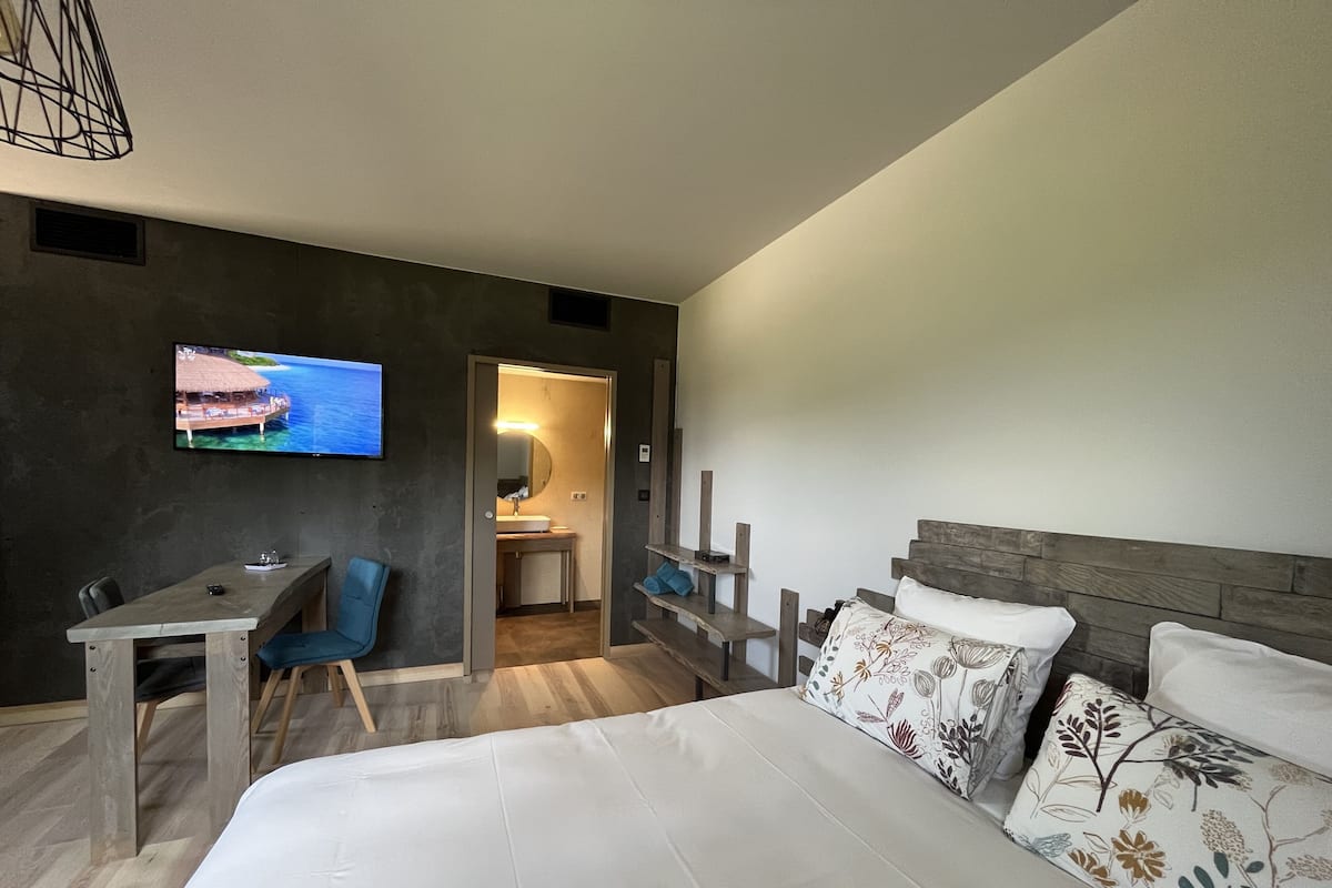 Une chambre d’hôtel moderne, dotée d’un grand lit, d’un bureau, d’une chaise, d’une télévision et d’une salle de bain visible à travers une porte ouverte.