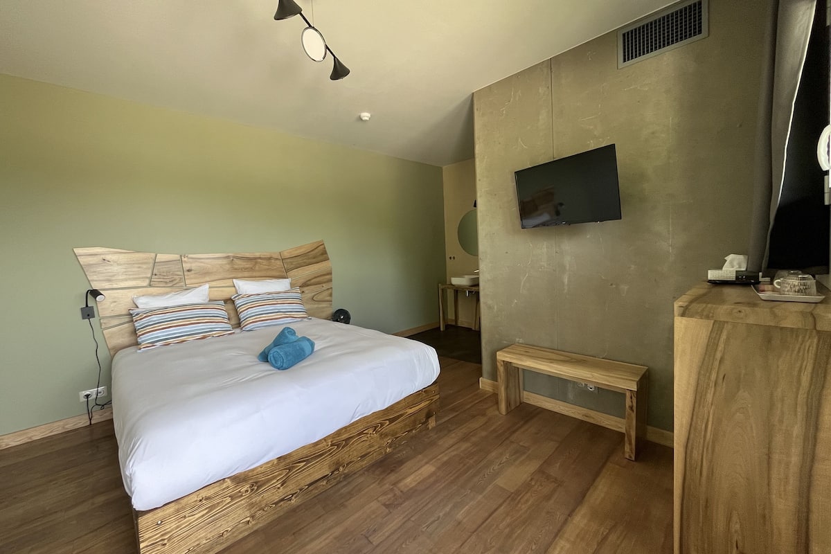 Une chambre avec une tête de lit en bois, un lit recouvert de draps blancs, une table de chevet, un petit banc en bois, un téléviseur à écran plat fixé au mur et une fenêtre donnant sur l’extérieur.