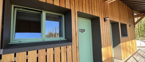 A building with wooden paneling, a green door labeled "Orme 5", and a window with a view of trees.