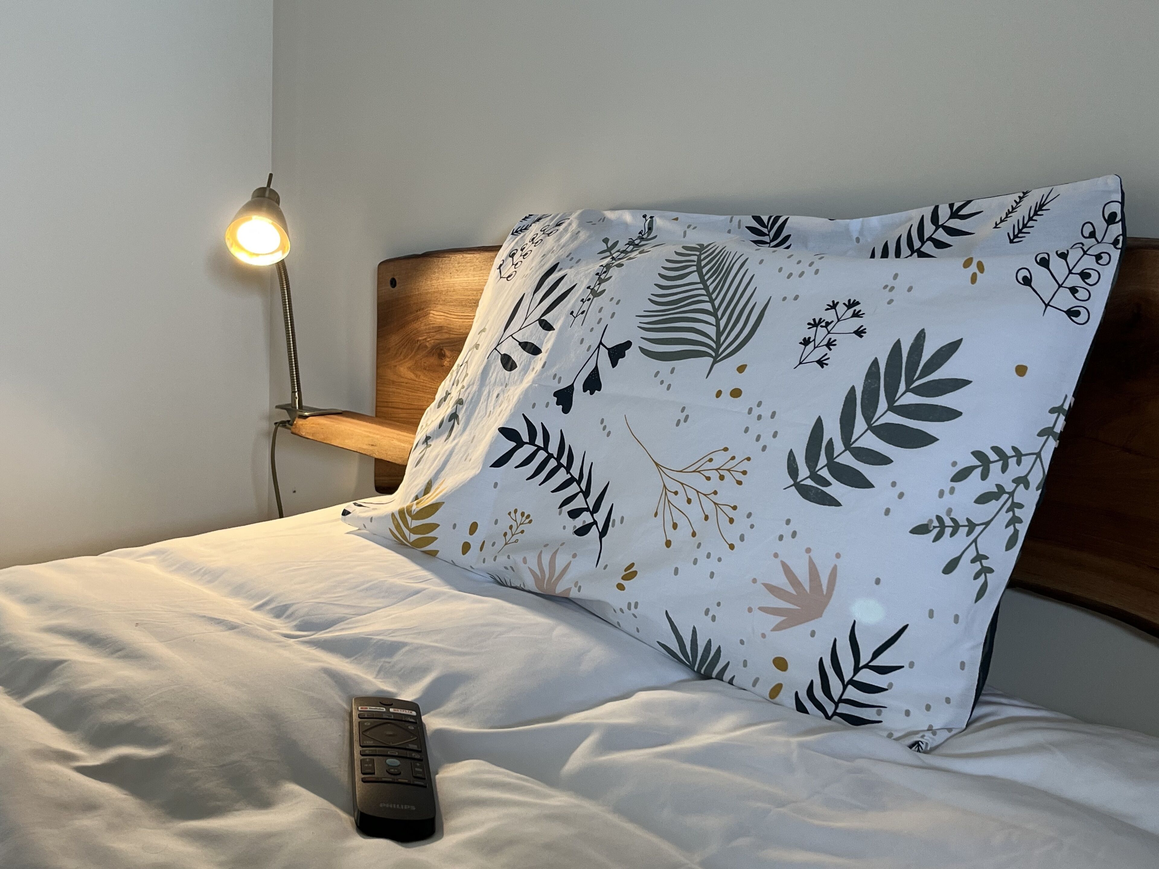 Een bed met een witte lakenset en een kussen met een bladmotief. Een afstandsbediening ligt op het bed.