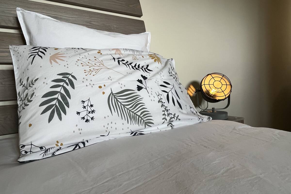 Un lit avec un drap blanc et un coussin décoratif orné d’un motif de feuilles. Une table de chevet avec une lampe se trouve à côté du lit.