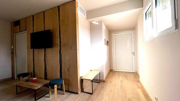 A room with a wooden wardrobe, a flat-screen TV mounted on the wall, a small table with a chair, and a door with a window.