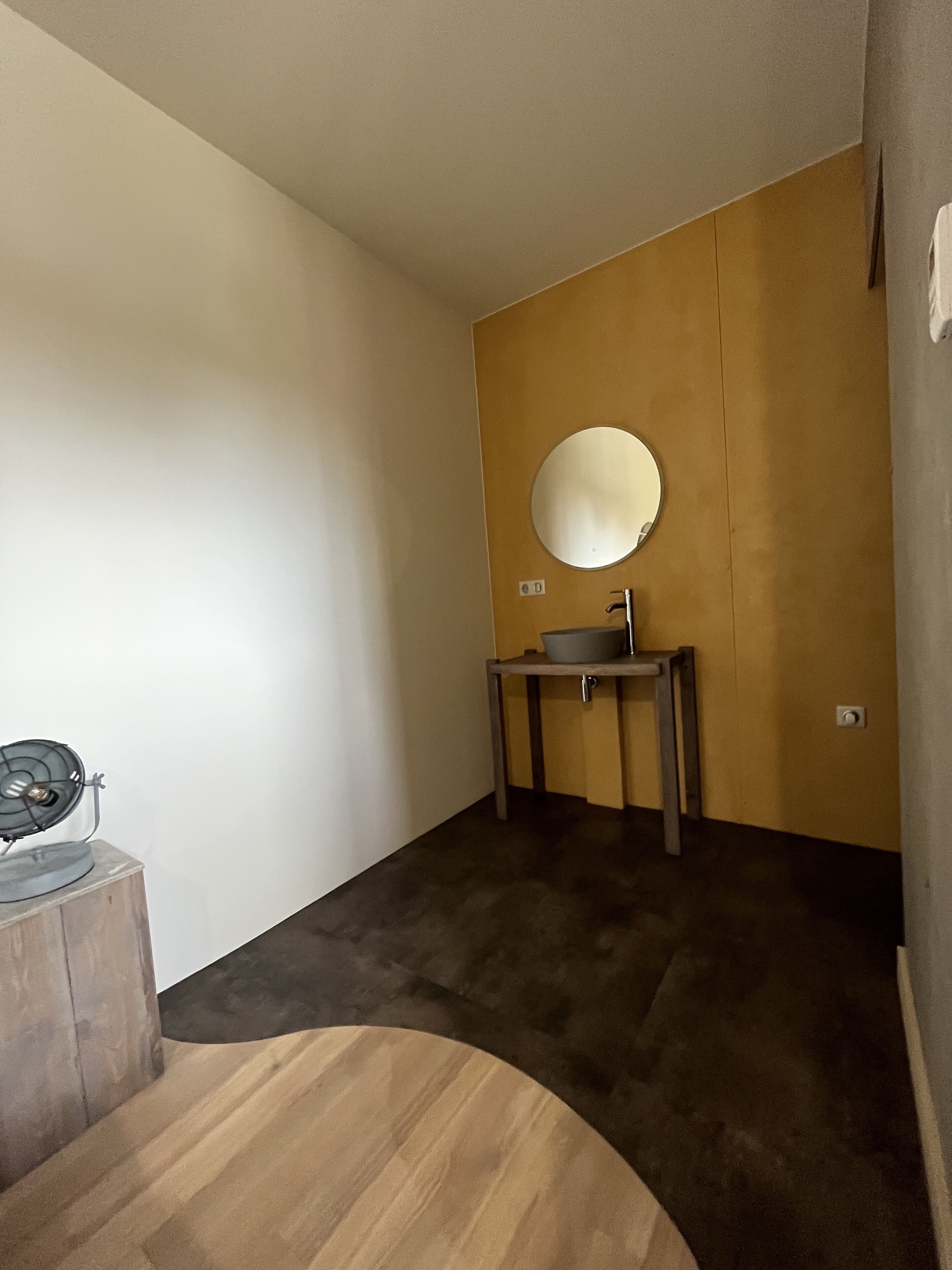 A small bathroom with a round mirror, a sink, and a fan on a wooden stool.