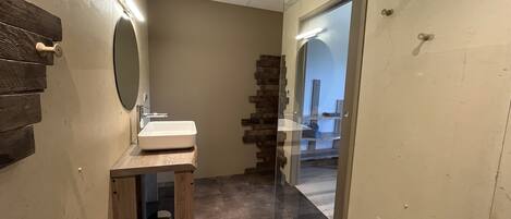 Un baño con tocador de madera, un lavamanos y un espejo. Hay una puerta de cristal que da a otra habitación.