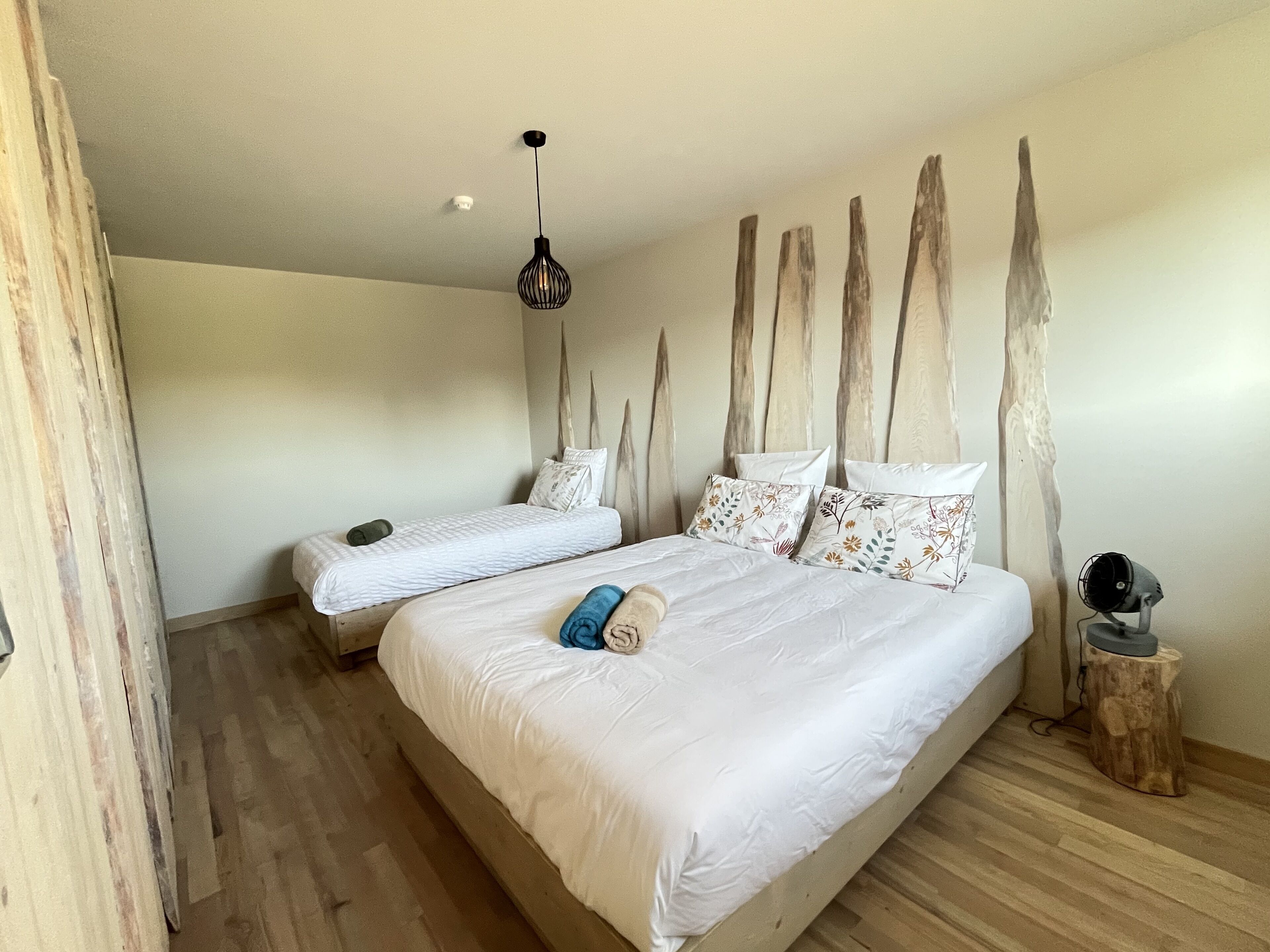 Un dormitorio con dos camas, piso de madera, lámpara colgante y un taburete pequeño de madera.