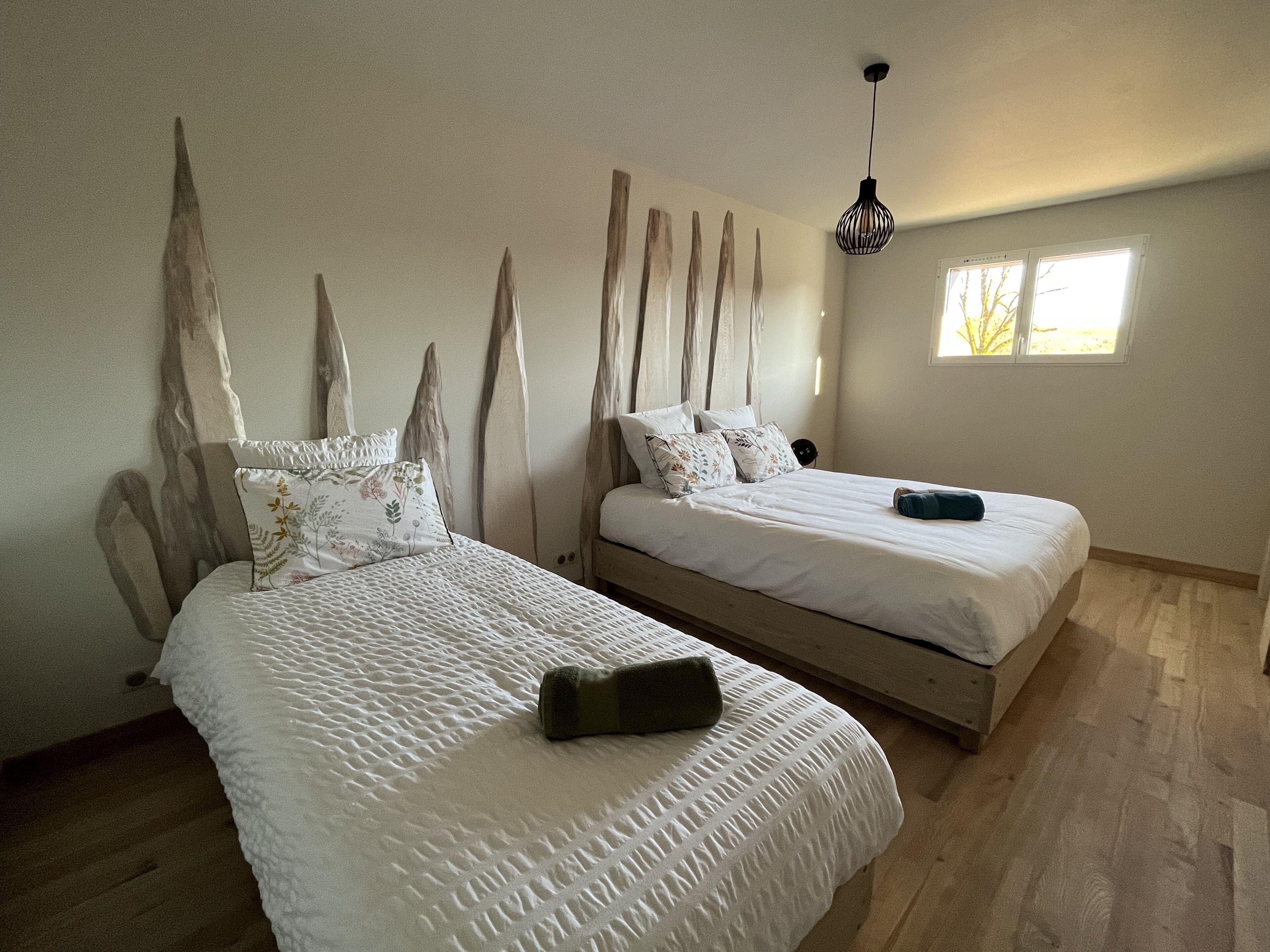 Un dormitorio con dos camas, cabeceros de madera, una lámpara colgante y una ventana con luz natural.