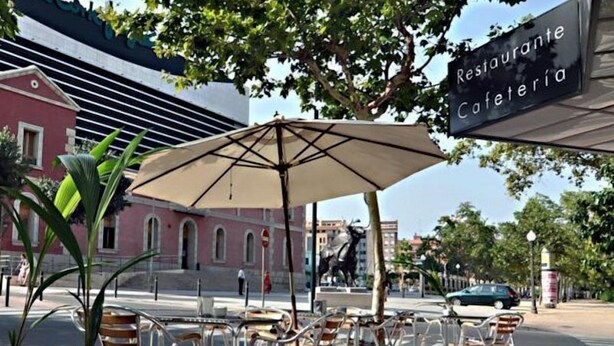 Mobiliario exterior con mesas y sillas, una sombrilla grande y un cartel de Restaurante Cafetería.
