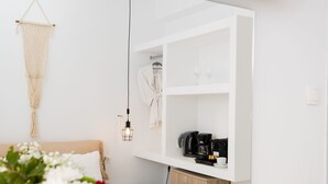 Een moderne, minimalistische kamer met een bed, een macramé wanddecoratie, een koffieapparaat en een wit schap met een badjas en brillen.