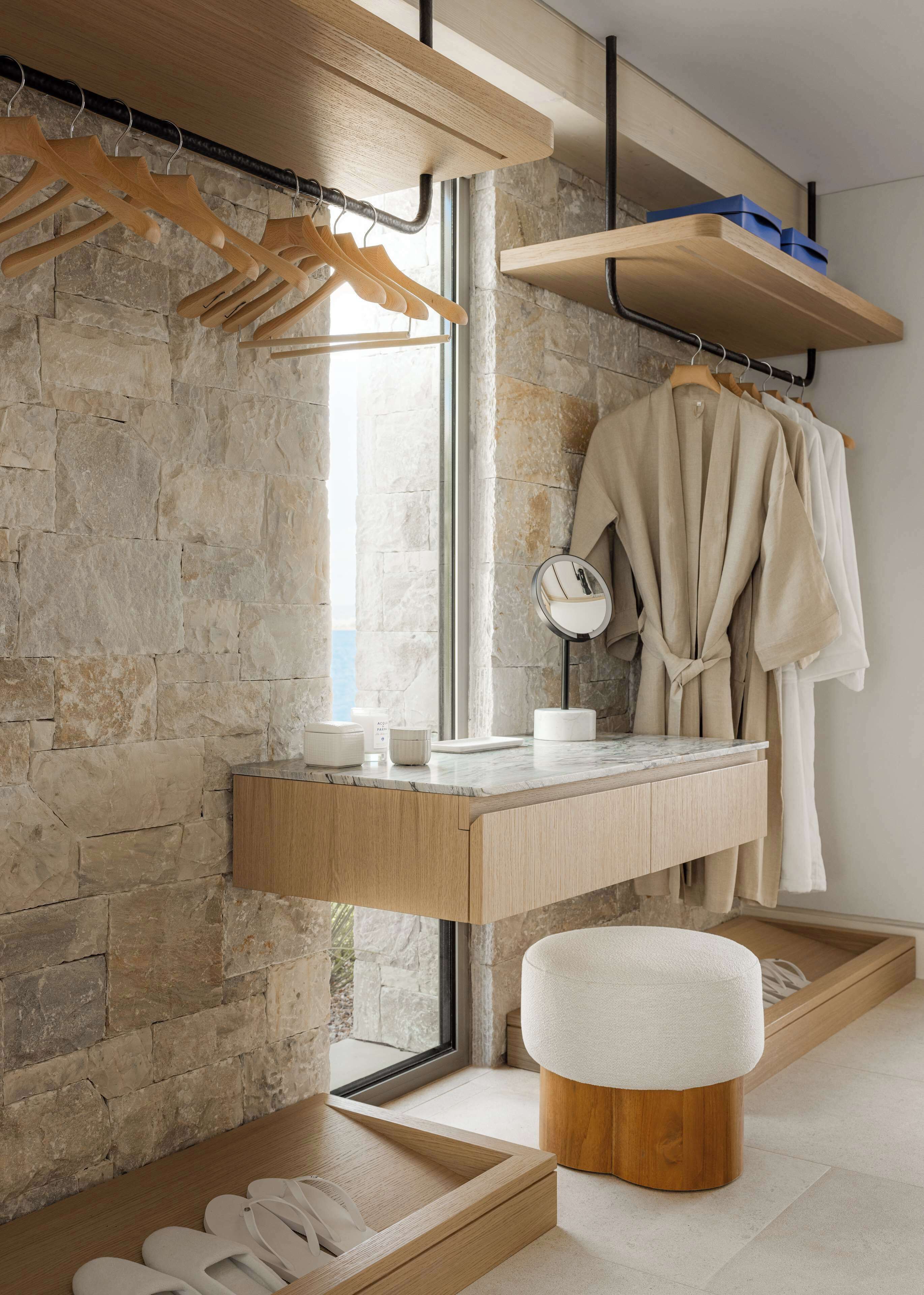 Een moderne badkamer met een stenen wand, houten wastafelmeubel, kapstok voor gewaden en een kleine ronde spiegel.