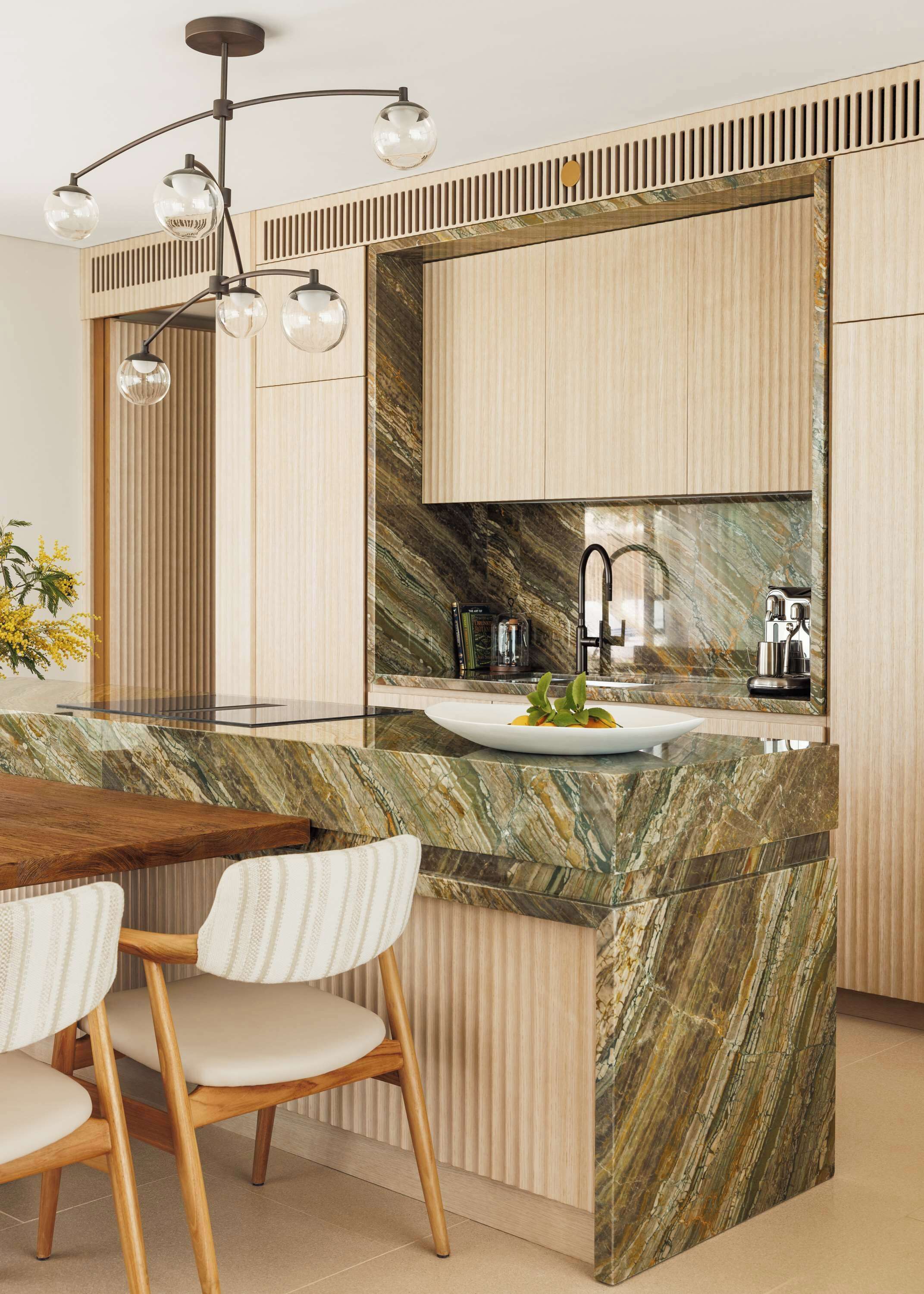 Una cocina moderna con una isla de mármol, sillas de madera, un lavamanos empotrado y una lámpara colgante.