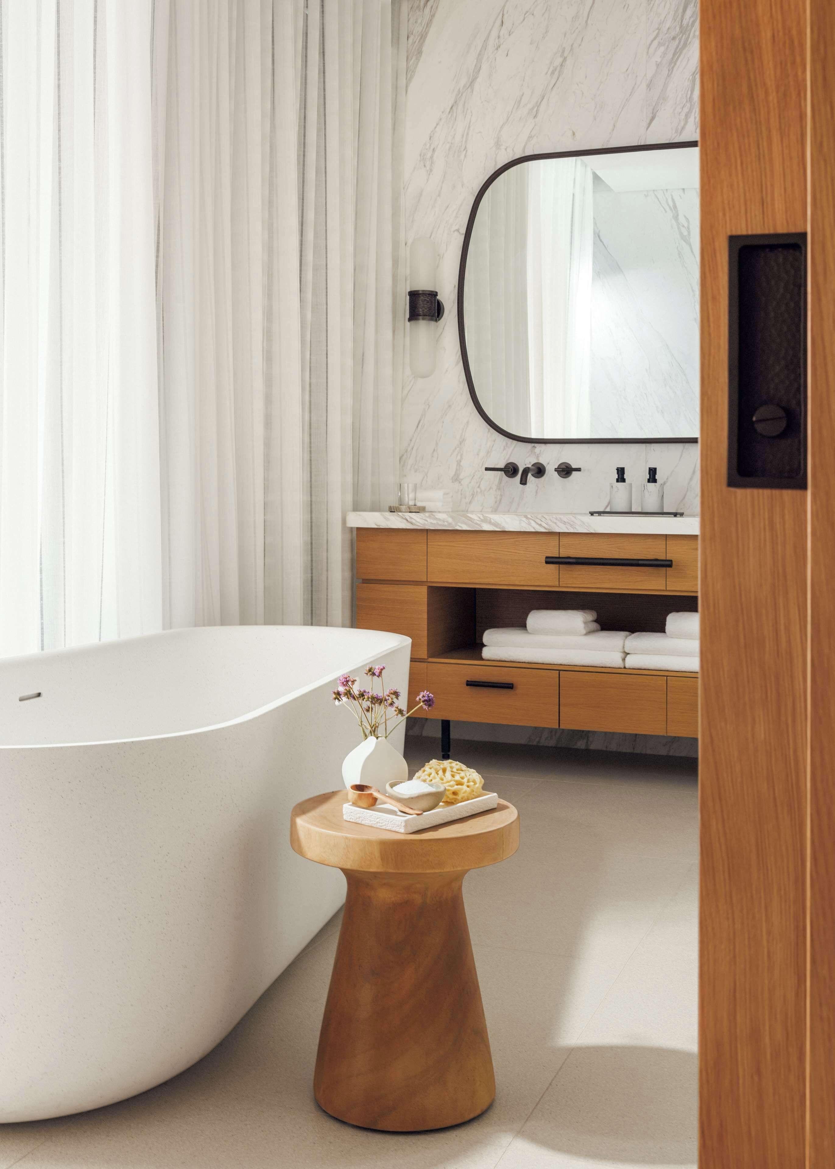 A modern bathroom with a freestanding bathtub, a wooden vanity with a marble countertop, and a round wooden side table with a vase and a bowl.