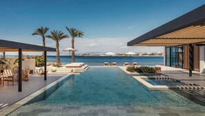 A modern infinity pool with clear blue water, surrounded by lounge chairs and palm trees, overlooking a scenic coastal view. - One&Only Aesthesis (Glyfada)