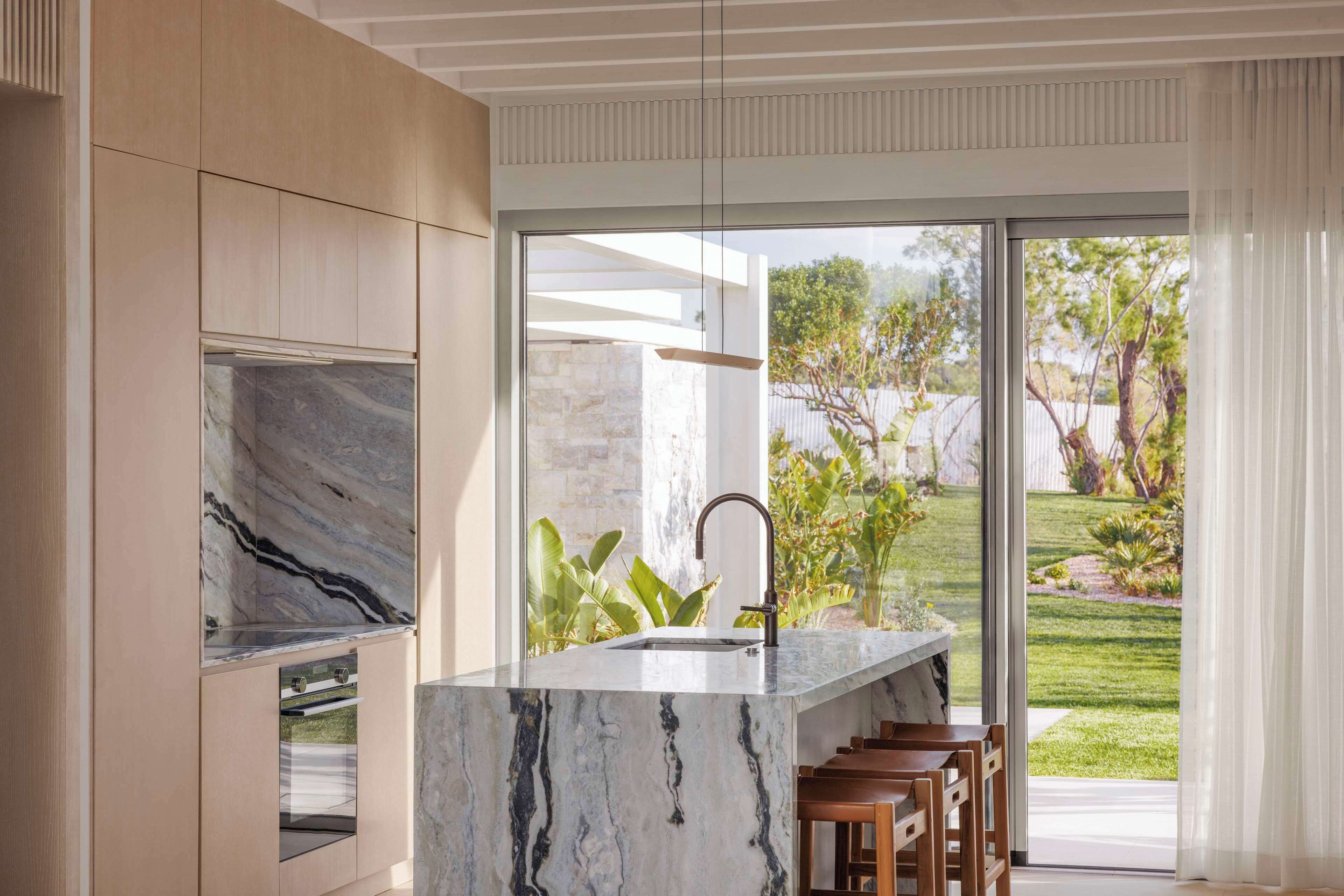A modern kitchen with a marble countertop, a sink, and a large window overlooking a garden.