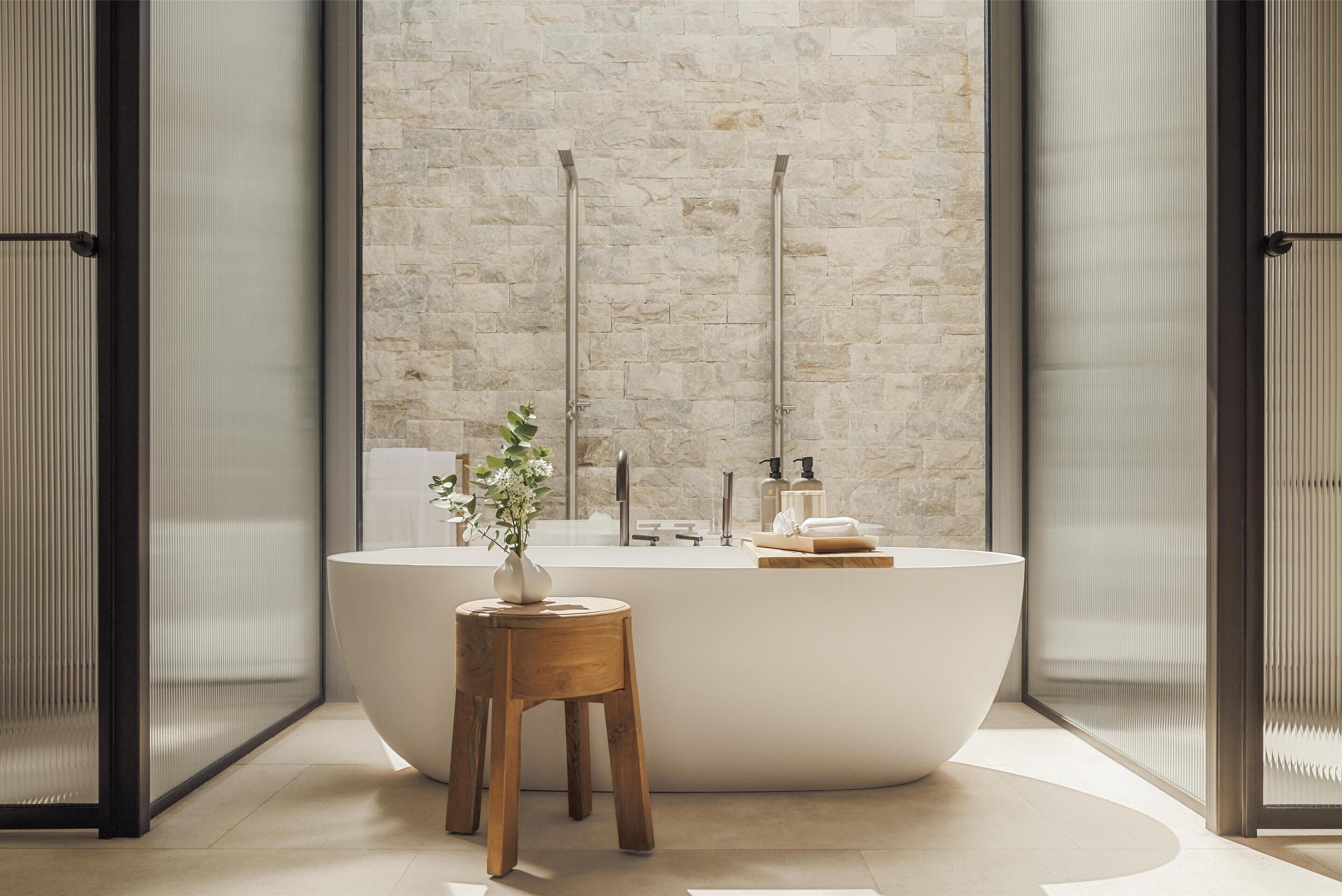A modern bathroom with a large freestanding bathtub, a wooden stool, and a vase of flowers.