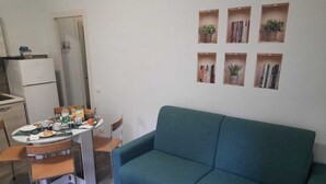 A compact living space with a dining table, chairs, a sofa, and a kitchenette. There are bookshelves with books and potted plants on the wall.