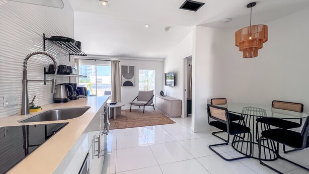 A modern kitchen with a large island, a dining area with a glass table and black chairs, and a hanging wooden light fixture.