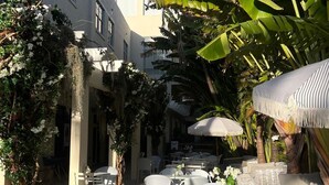 An outdoor café area with round tables and white wicker chairs, surrounded by lush greenery and a white building with balconies.
