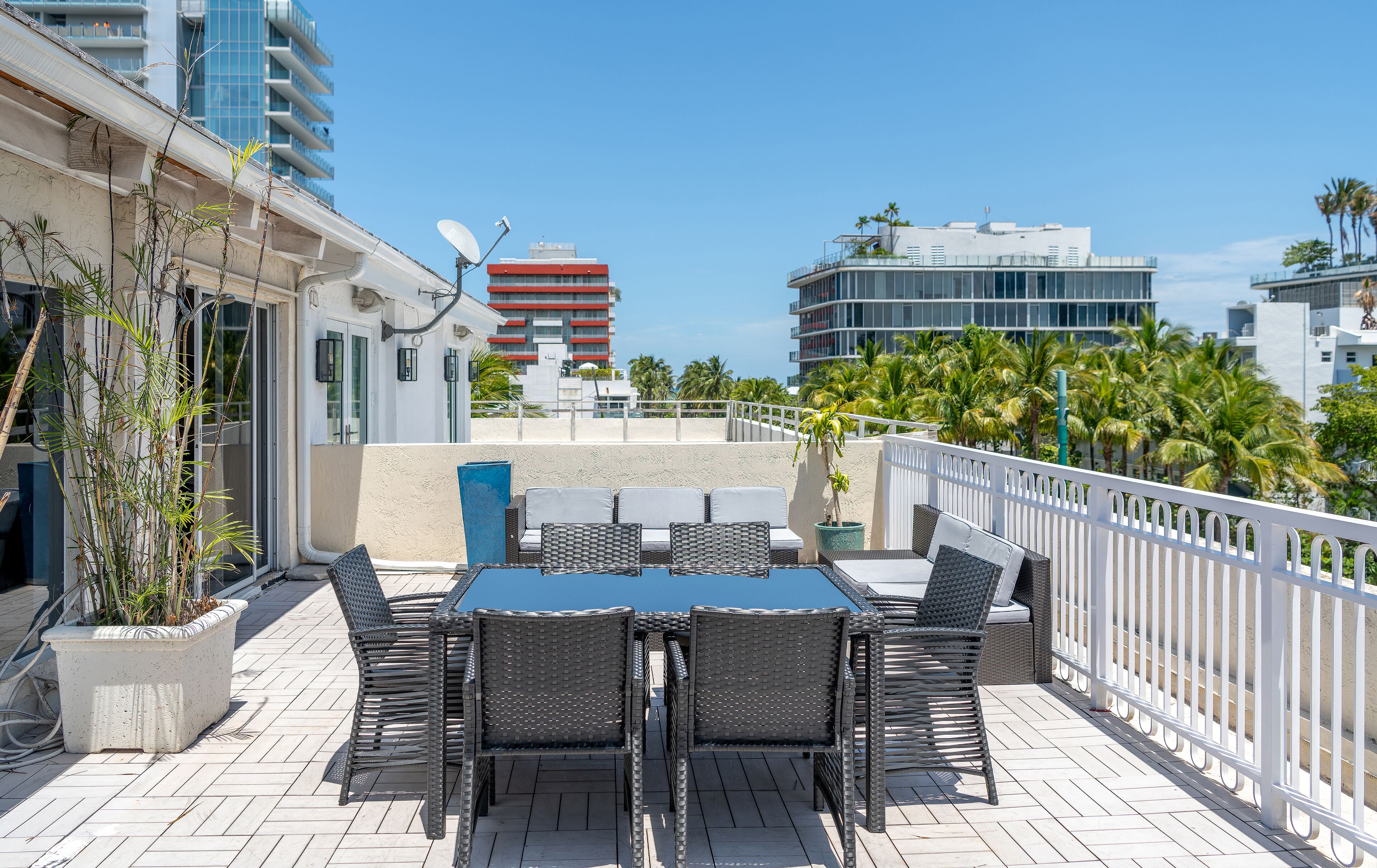 Een terras buiten met een tafel en stoelen, een witte balustrade en een uitzicht op palmbomen en gebouwen op de achtergrond.