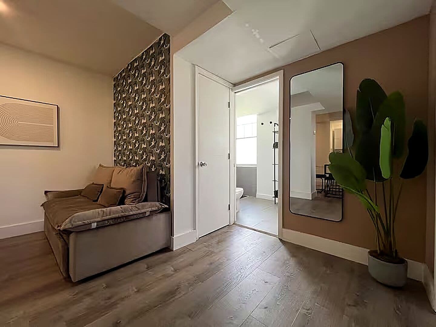 Una habitación con un sofá, un espejo, una planta en maceta y una puerta que da a otra habitación.