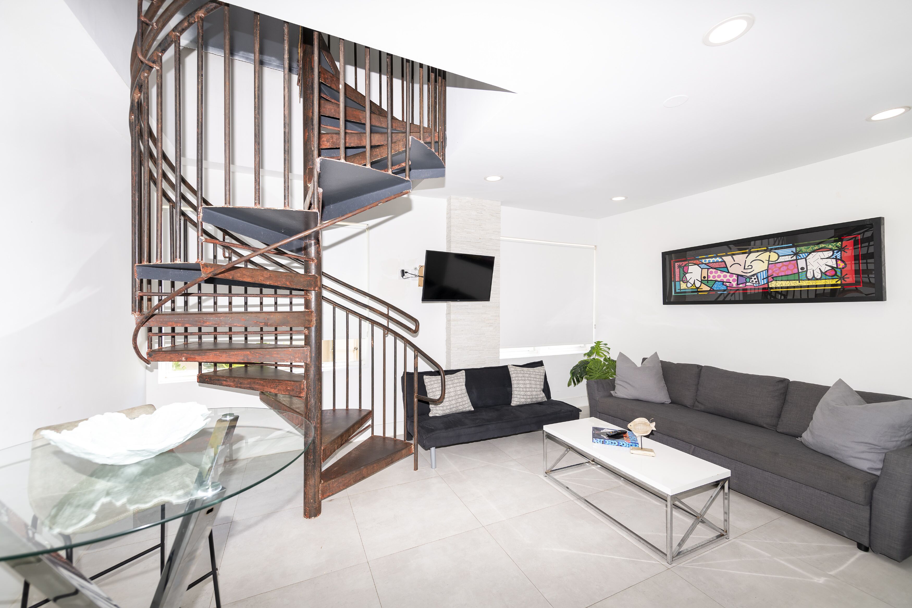 Un salon moderne avec un escalier en colimaçon, un téléviseur à écran plat, un canapé gris et une table basse en verre.