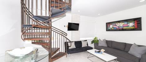 Un salon moderne avec un escalier en colimaçon, un téléviseur à écran plat, un canapé gris et une table basse en verre.