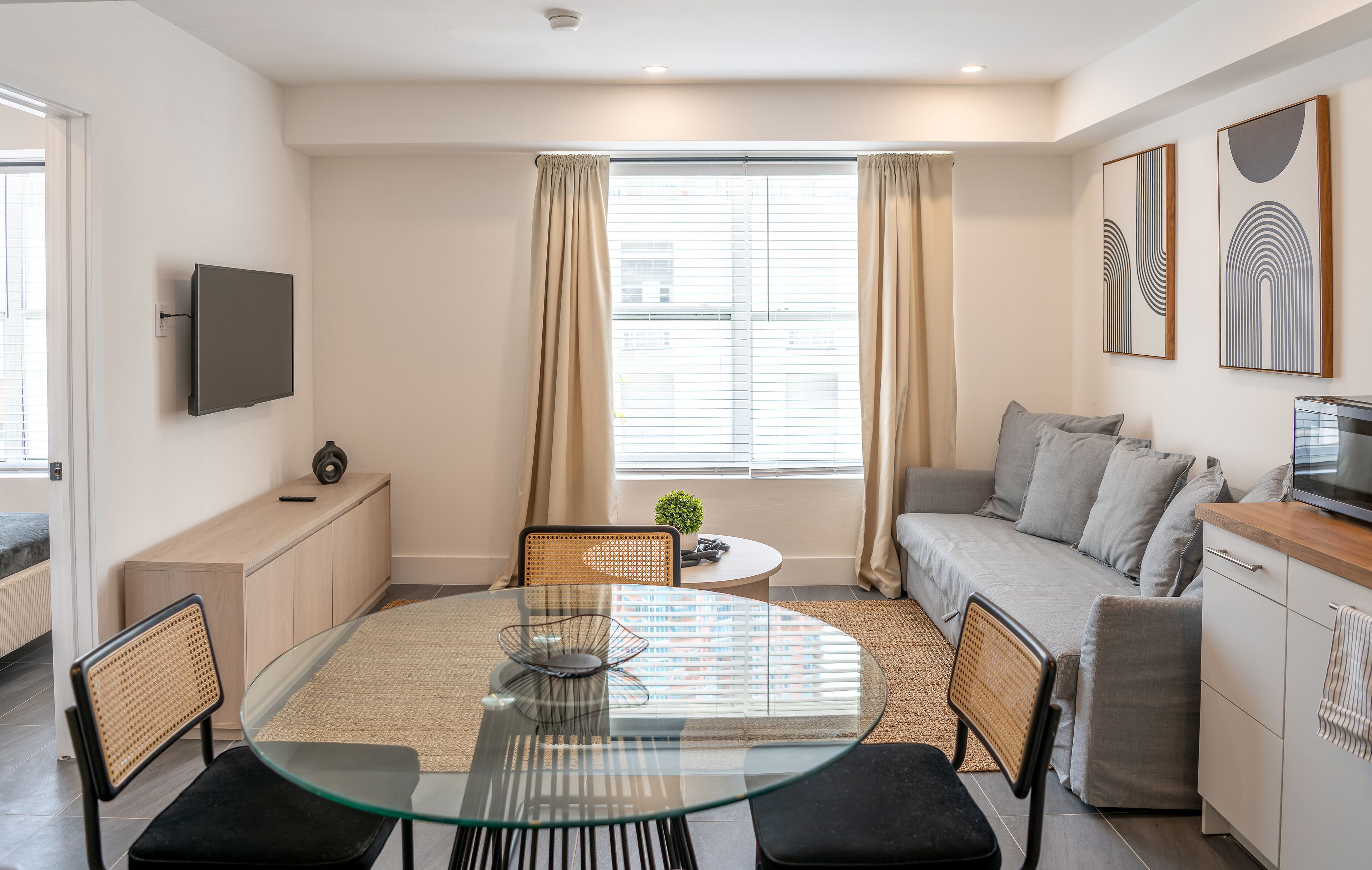 A modern living room with a glass dining table, a sofa, and a TV mounted on the wall.