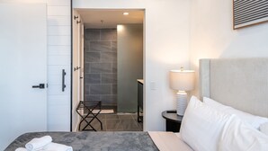 A hotel room with a bed, a nightstand, a lamp, a towel on the bed, and a shower area visible through an open door.