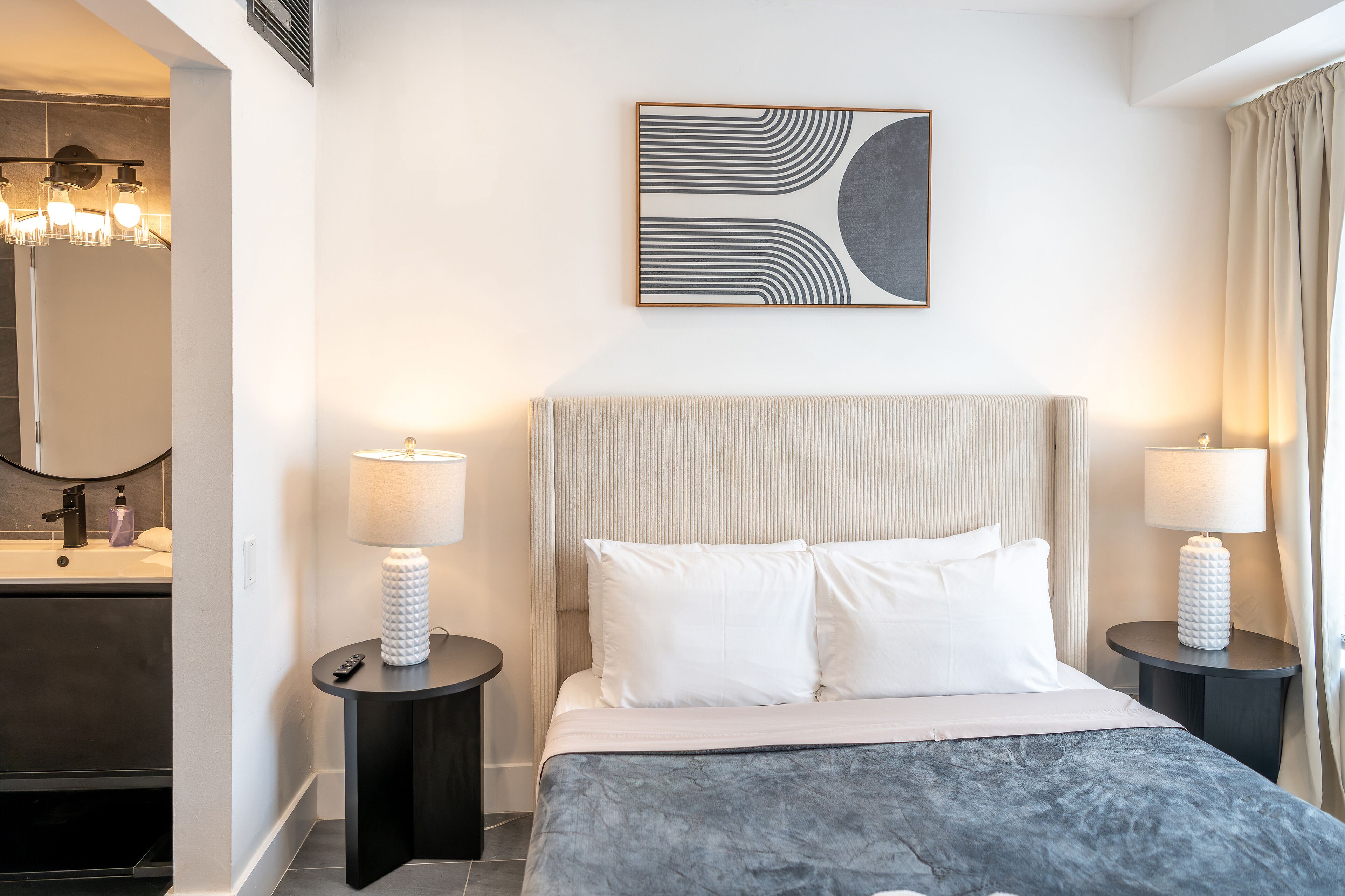 A modern hotel room with a large bed, two bedside tables with lamps, a bathroom with a mirror and sink, and a wall-mounted abstract art piece.