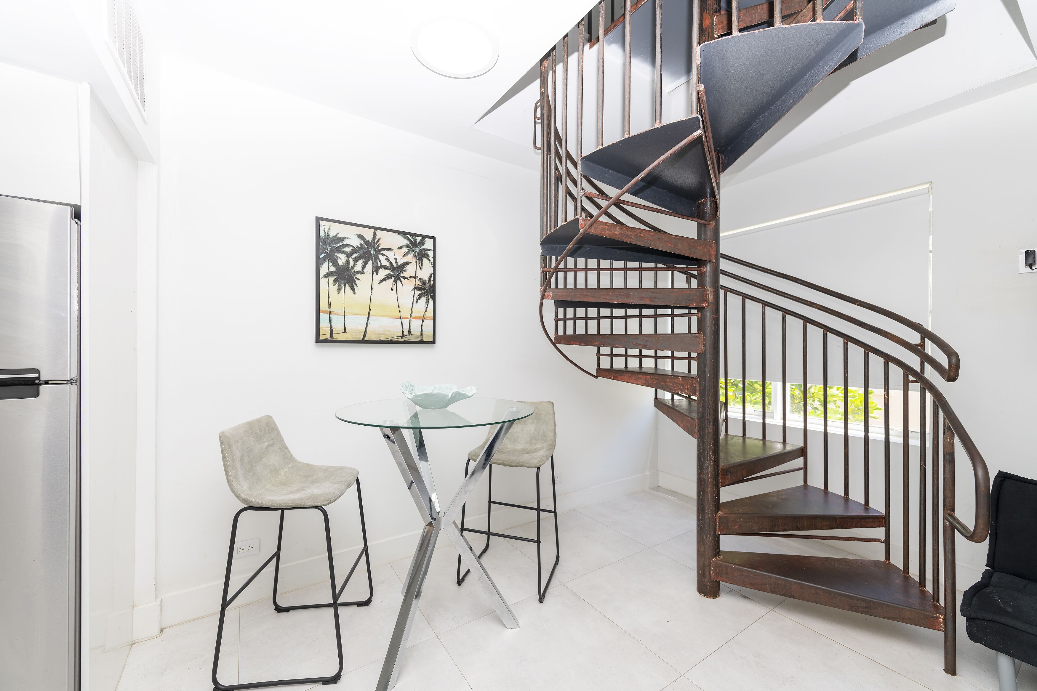 A modern kitchen with a stainless steel refrigerator, a glass table with two chairs, and a spiral staircase.