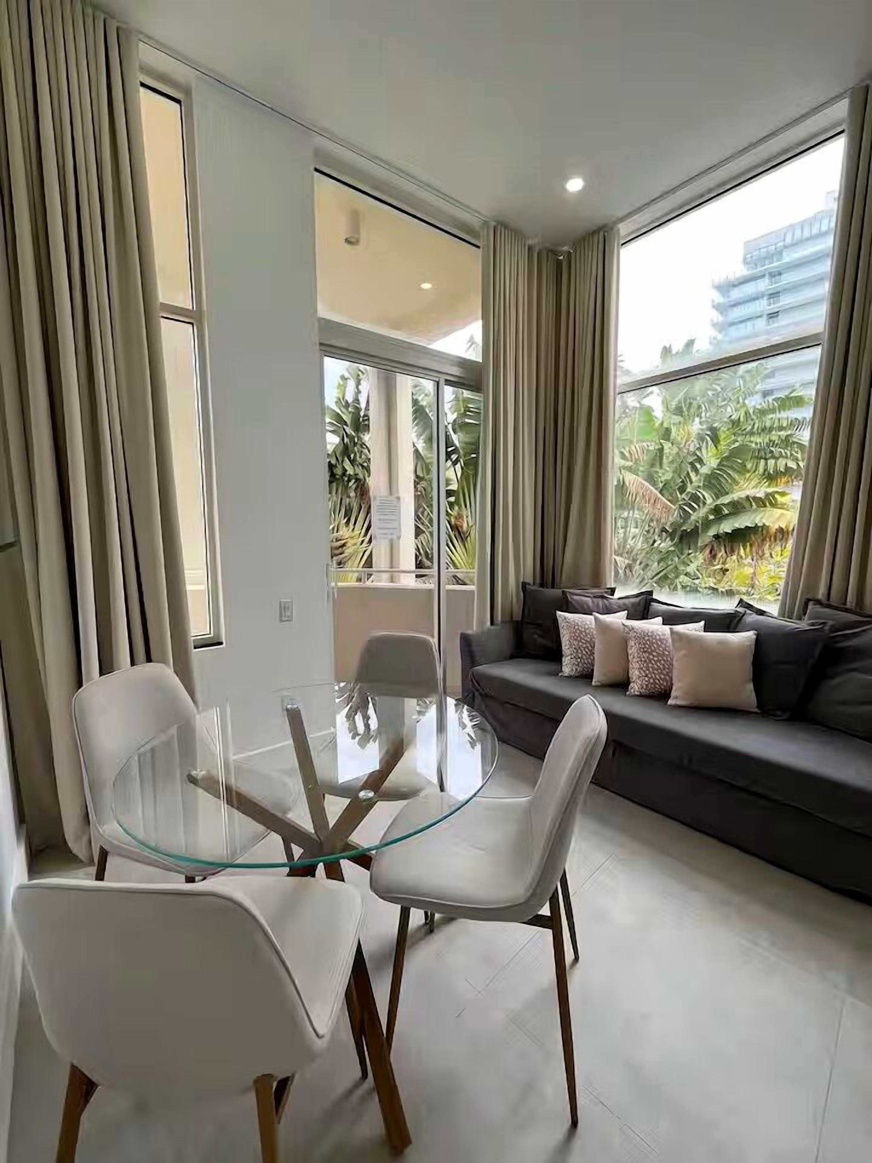A modern living room with a glass dining table, white chairs, a dark sofa with patterned cushions, and large windows offering a view of greenery and buildings.
