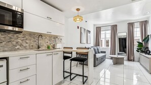A modern kitchen with white cabinets, a microwave, and a tiled backsplash. There is a dining area with a table and chairs, and a gray sofa.
