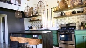 Une cuisine dotée d’un îlot central, de tabourets de bar, de luminaires suspendus, d’une cuisinière et d’étagères remplies d’ustensiles de cuisine.