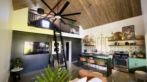 Un vaste espace de vie doté d’un ventilateur de plafond, un coin repas avec des chaises, une cuisine équipée d’une cuisinière et un escalier menant à un niveau supérieur.