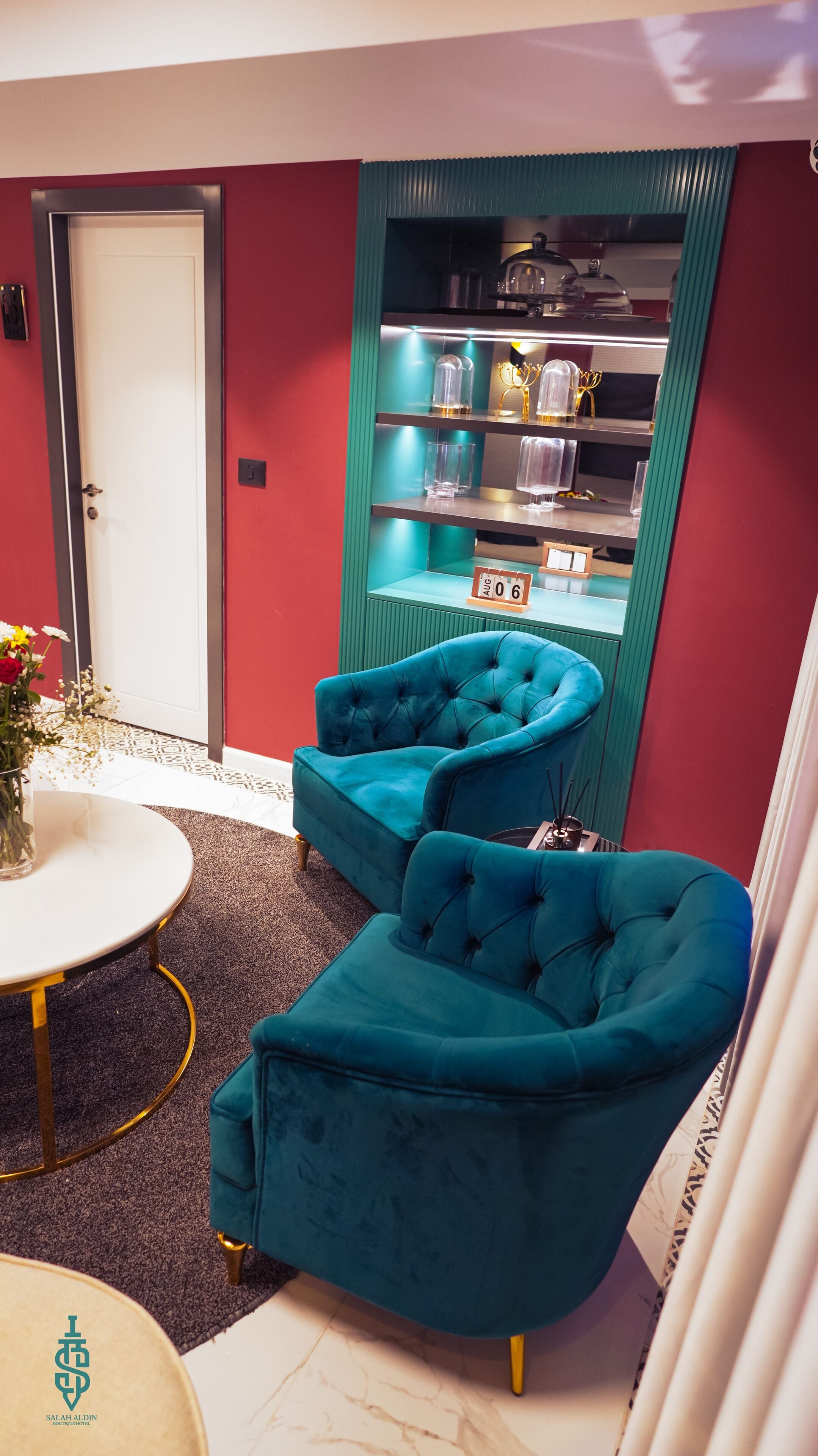 A room with teal armchairs, a white table, and a green shelf with decorative items.