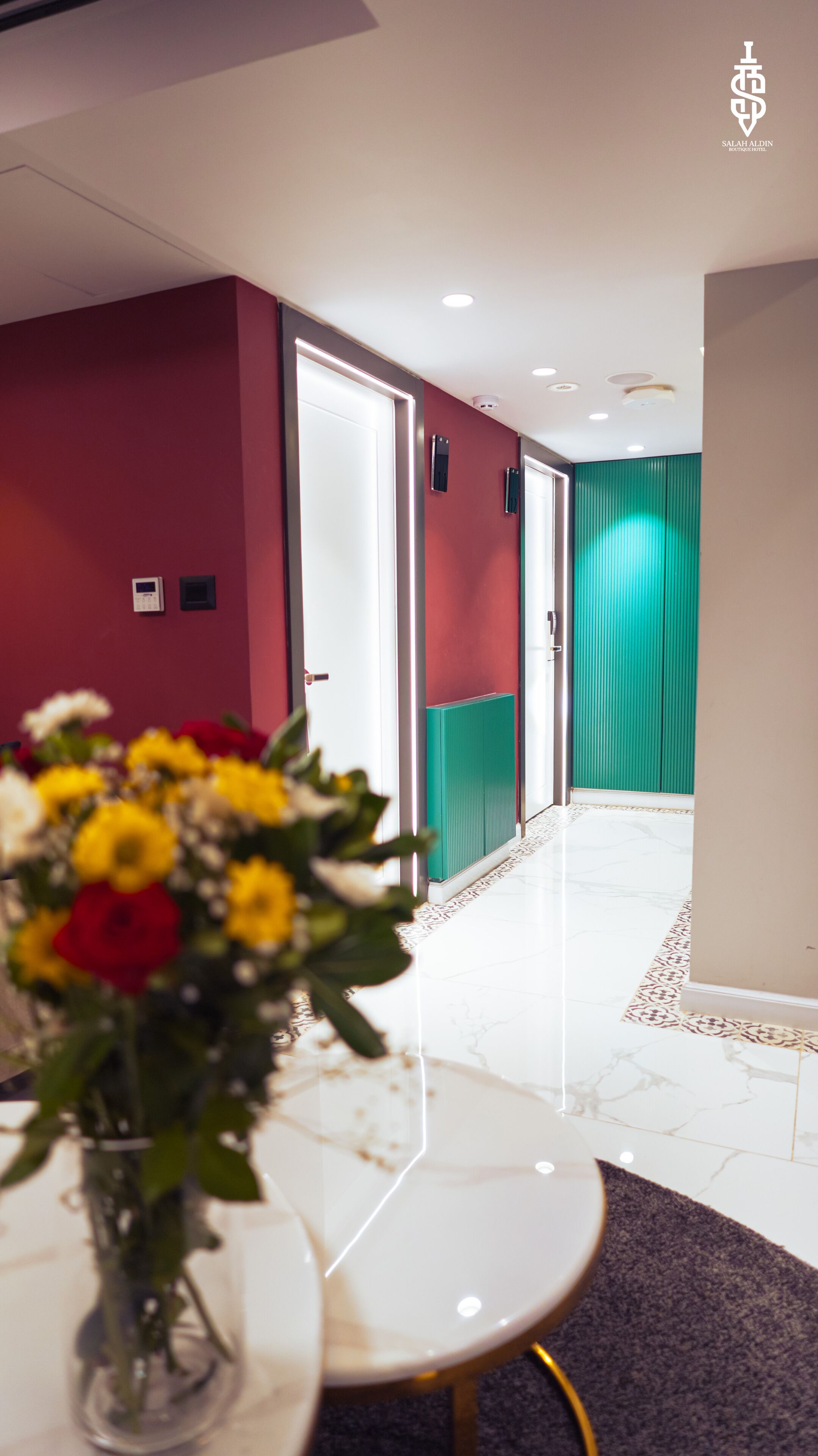 Um saguão de hotel com uma recepção de cor branca, um vaso com flores amarelas e vermelhas, uma parede vermelha e uma parede verde decorativa.