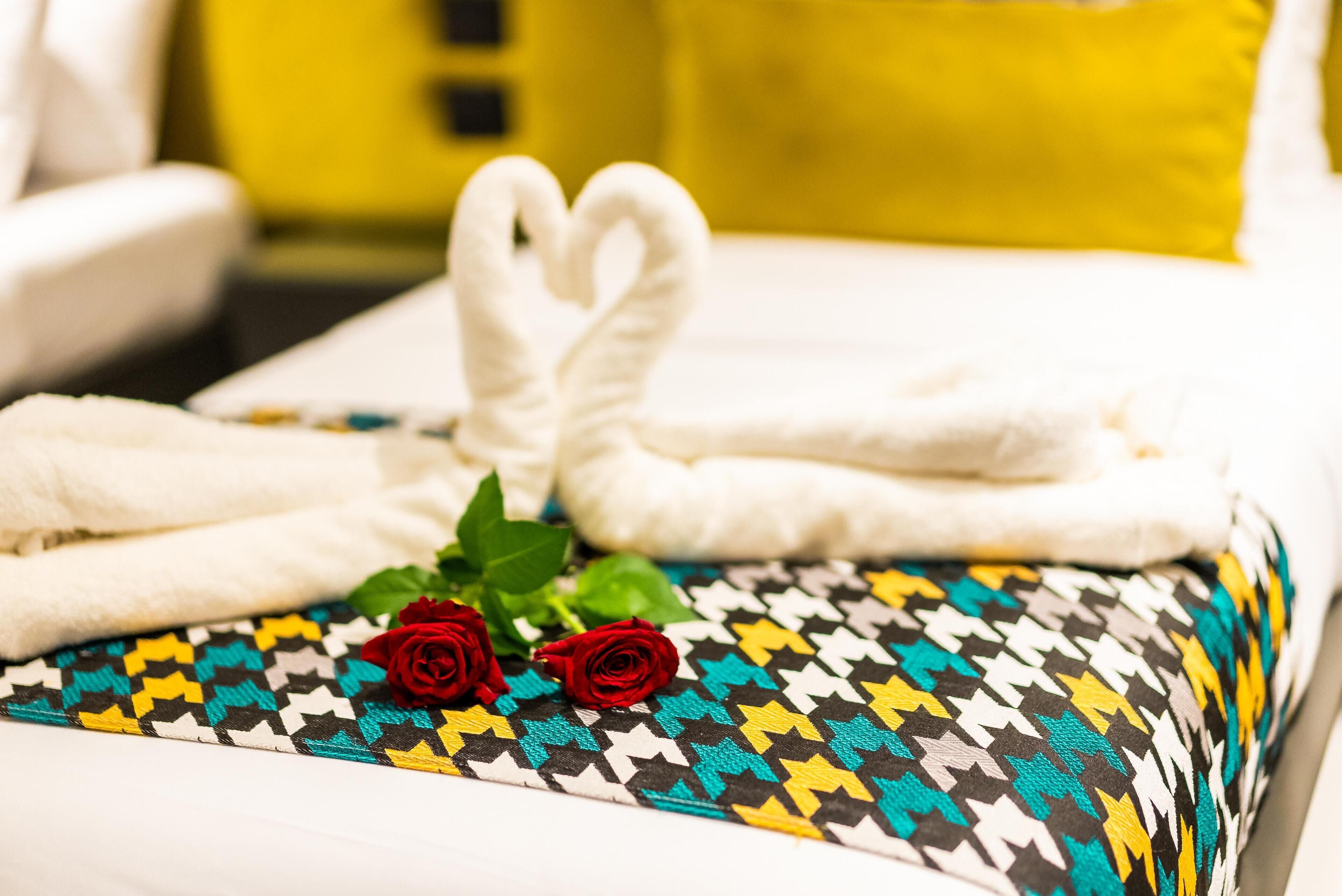 Cama arrumada com toalhas brancas dobradas em formato de coração, duas rosas vermelhas e almofada estampada.