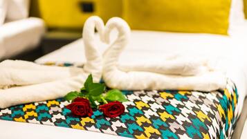 Cama arrumada com toalhas brancas dobradas em formato de coração, duas rosas vermelhas e almofada estampada.