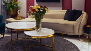 Un salon moderne avec un canapé beige, deux tables rondes à cadre doré, un tapis gris foncé, une plante en pot et un téléviseur à écran plat fixé au mur.