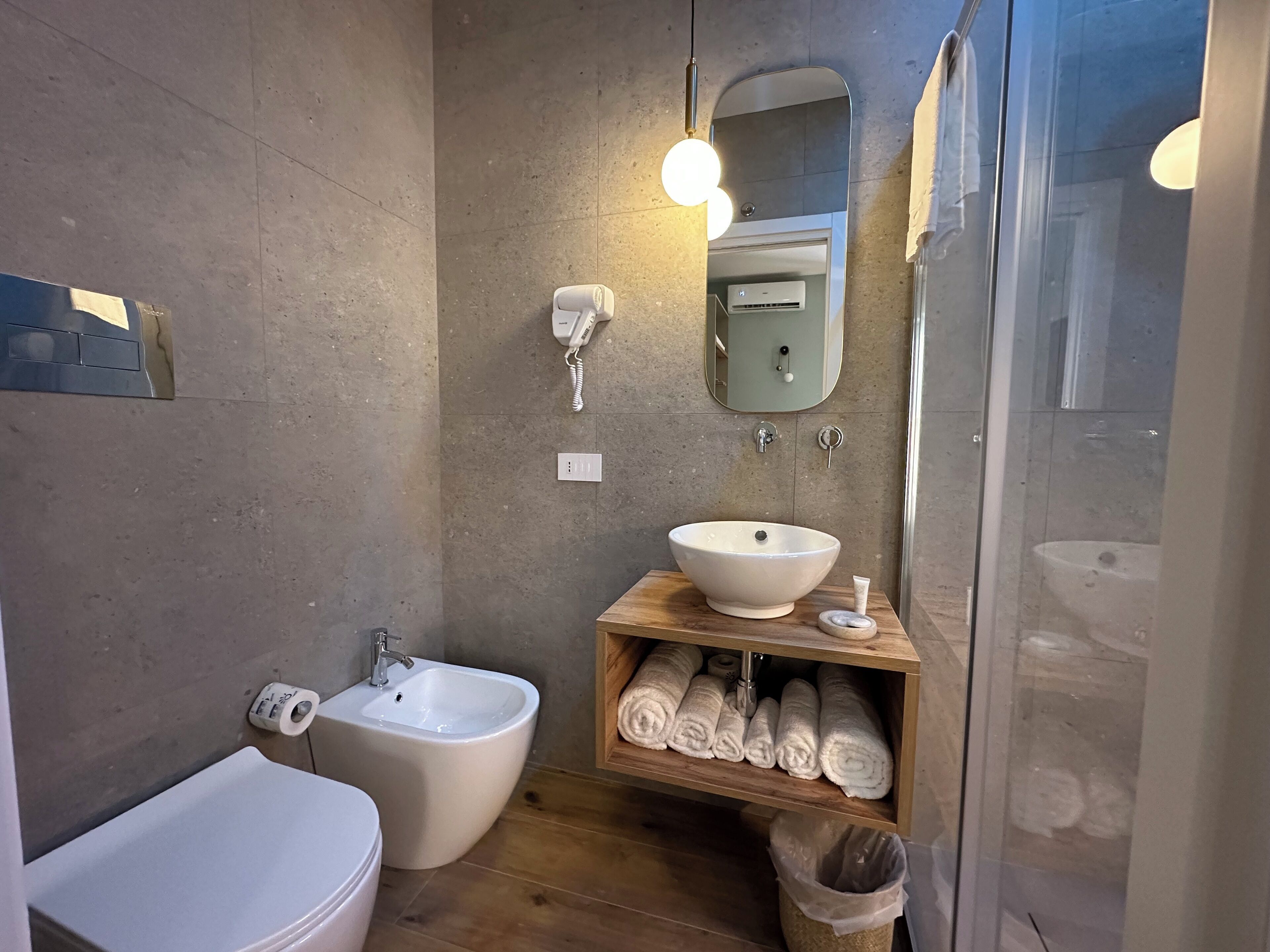 Un baño moderno con bidé, lavabo y espejo. Hay toallas en un estante y un cubo de basura.
