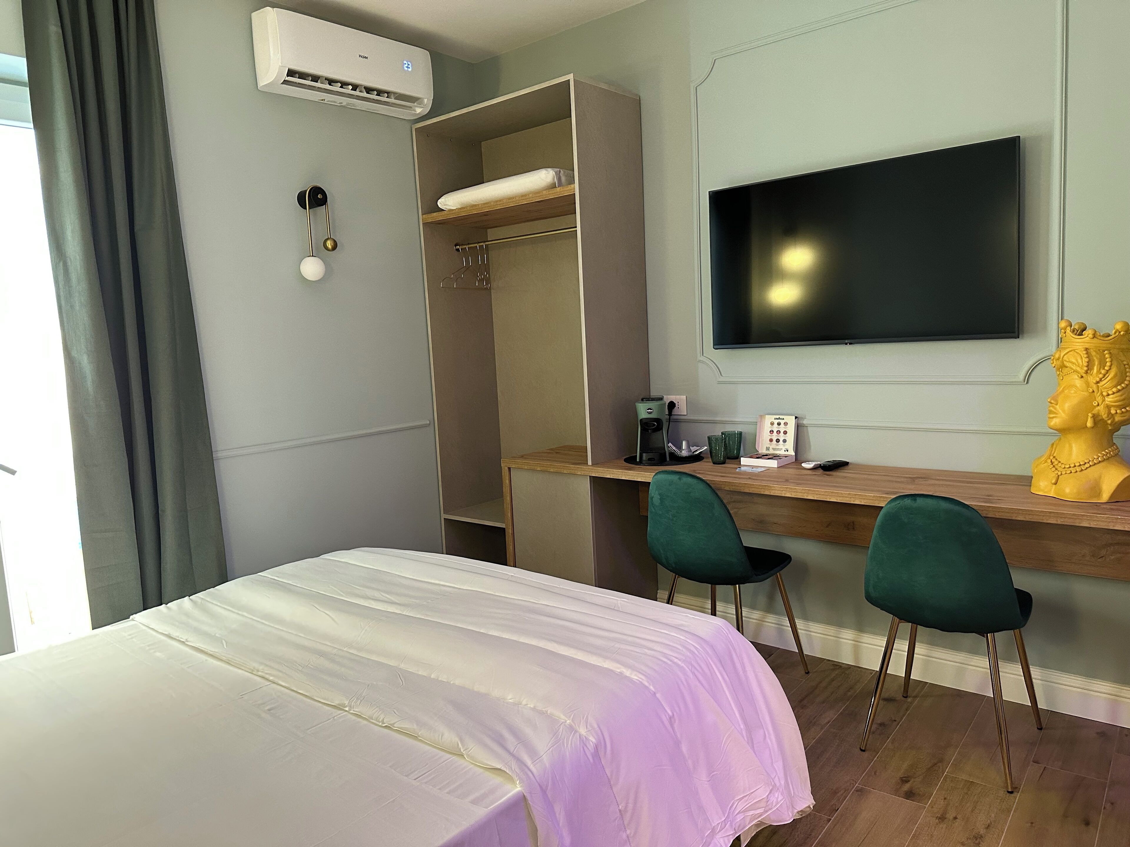 A hotel room with a bed, a desk with a chair, a TV, an air conditioner, and a wardrobe.