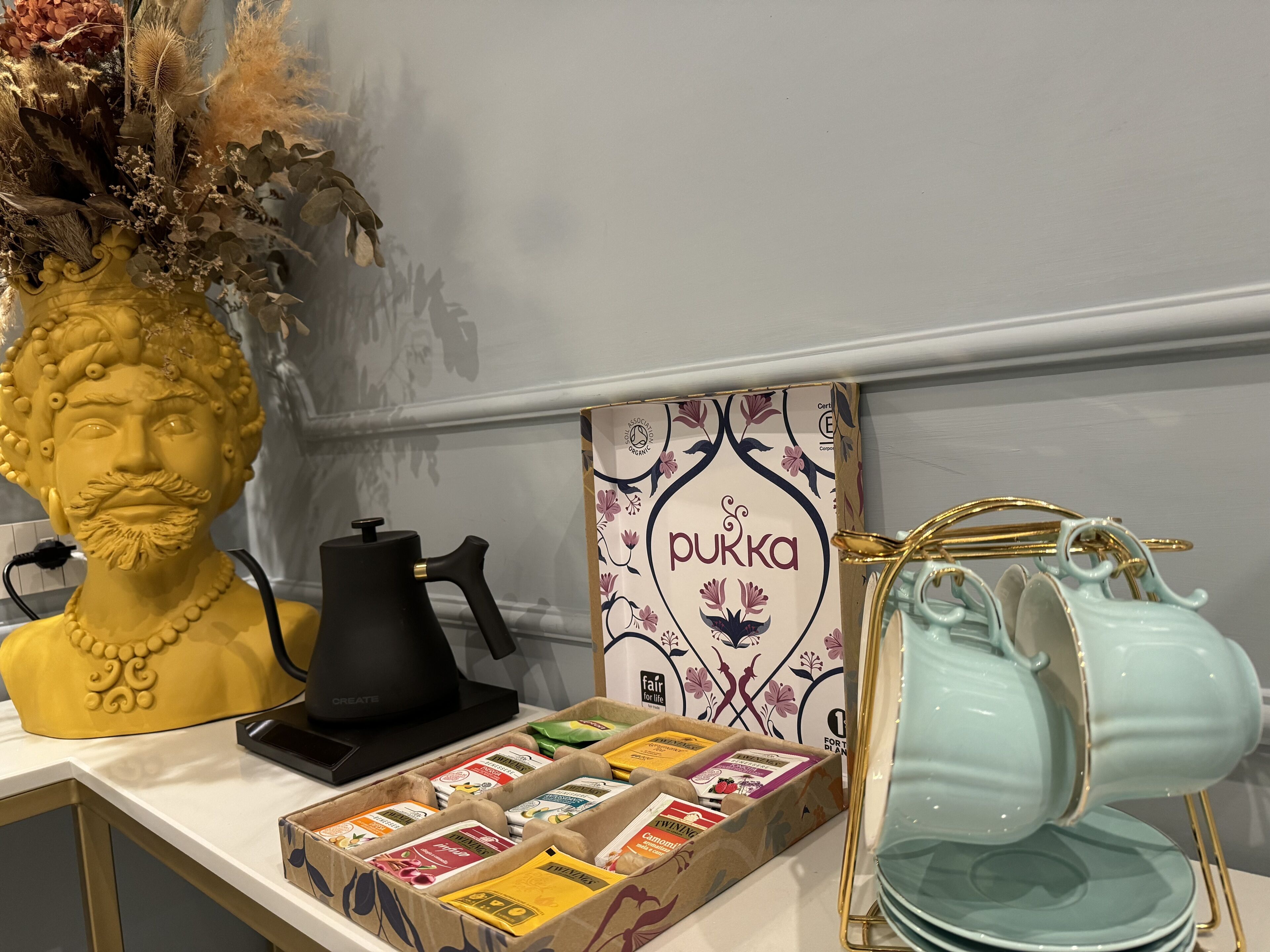 A table with a decorative bust, a black coffee maker, a box of tea bags, and a set of stacked plates.