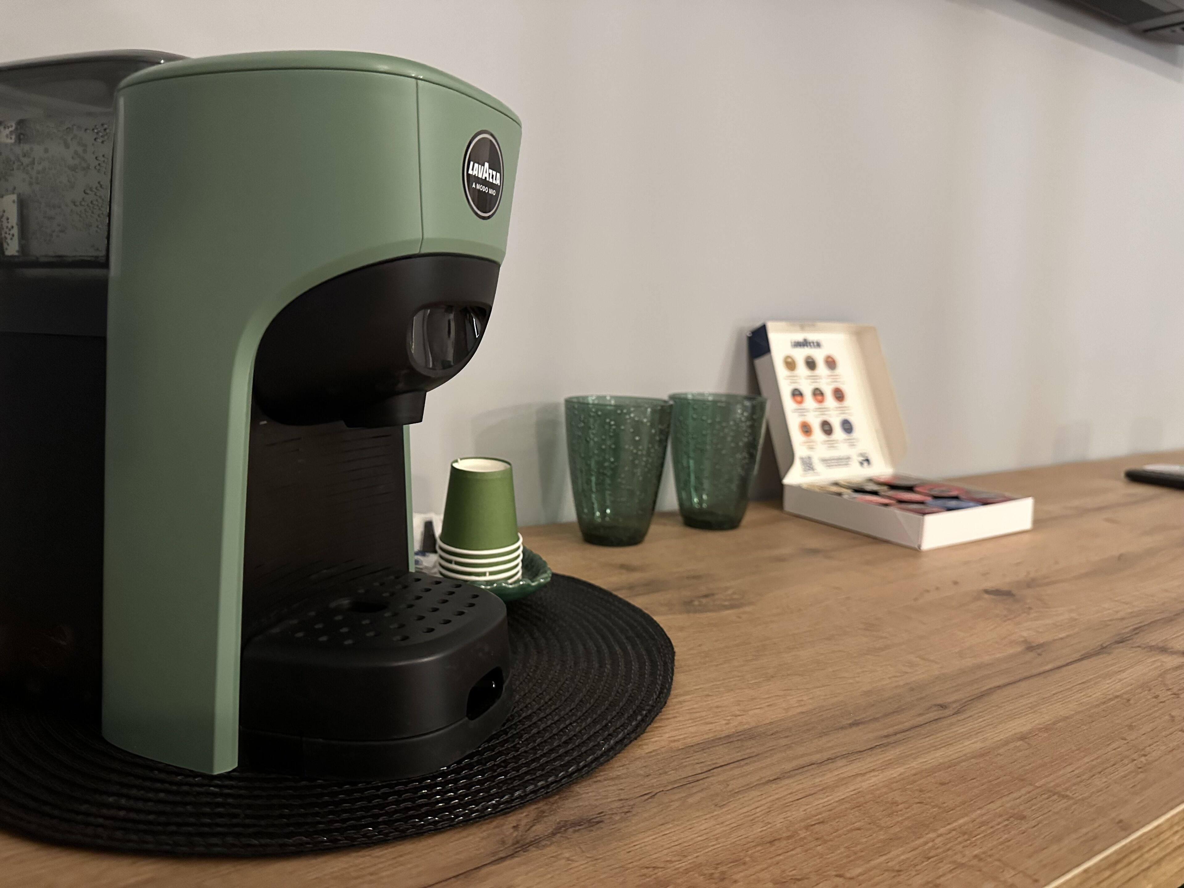 A coffee maker on a wooden table with a placemat, two green cups, and a coffee pod box.
