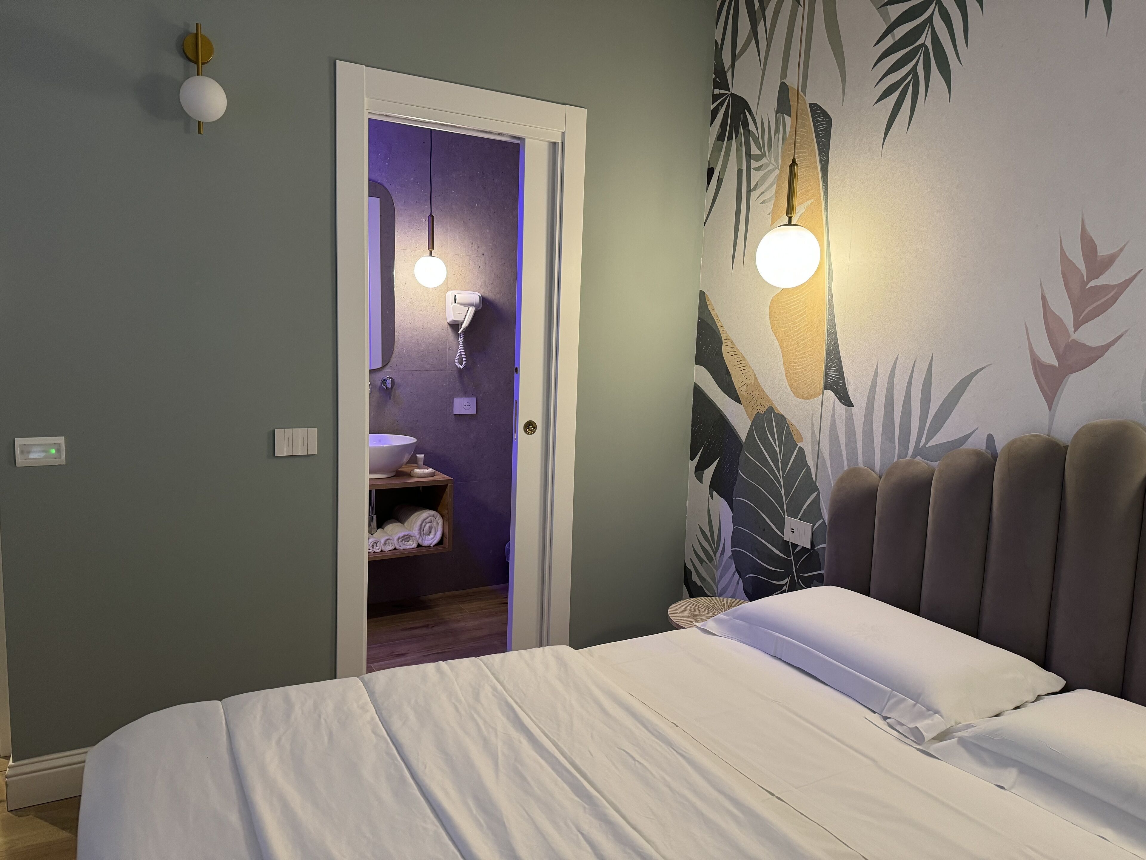 Un dormitorio con cama, mesita de noche, lámpara de pared, baño con lavamanos y toallero, y una puerta que da a otra habitación.