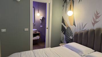 A bedroom with a bed, a nightstand, a wall-mounted lamp, a bathroom with a sink and towel rack, and a door leading to another room.