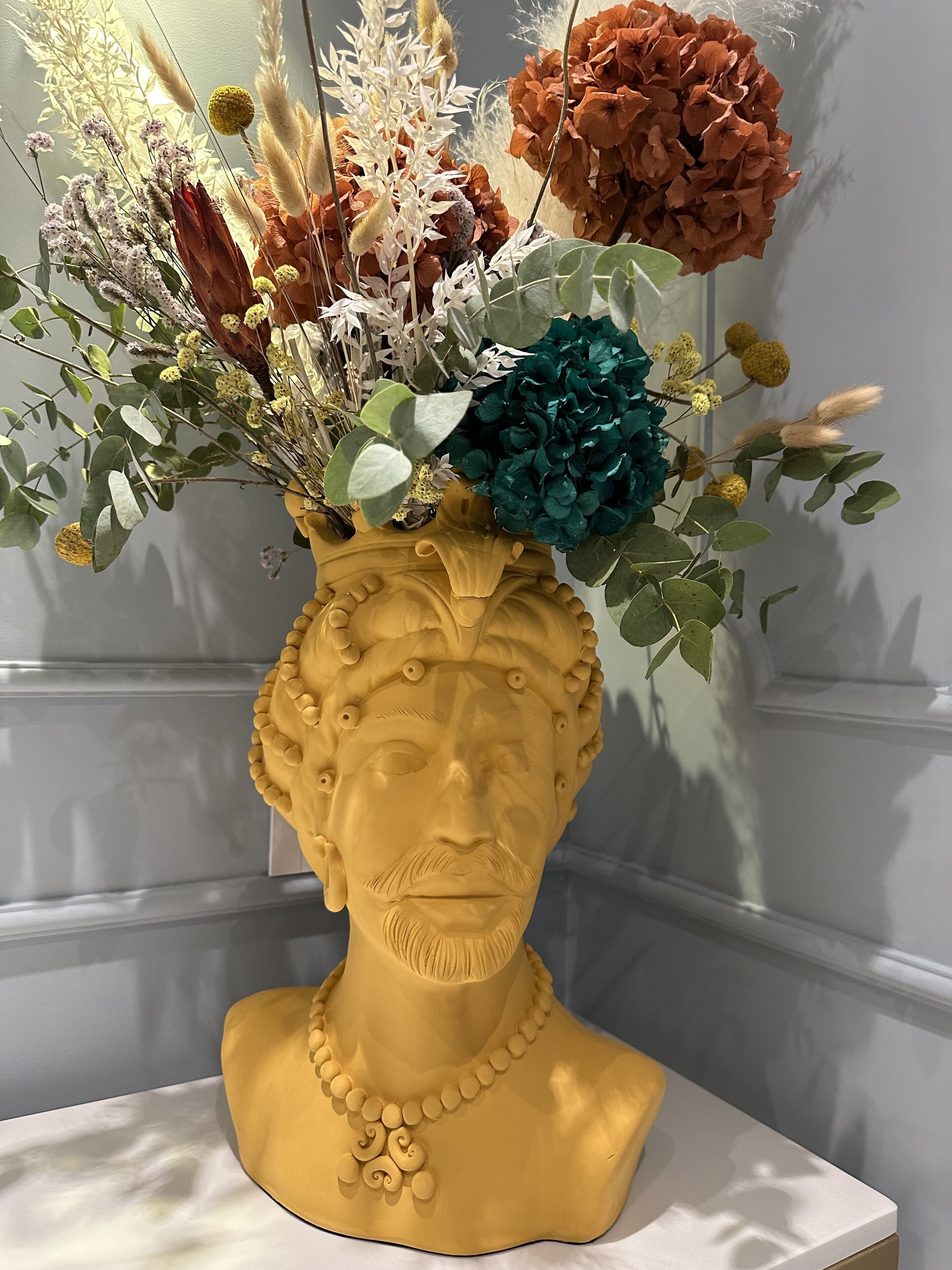 Escultura de busto con un arreglo floral en la parte superior, frente a una pared lisa.