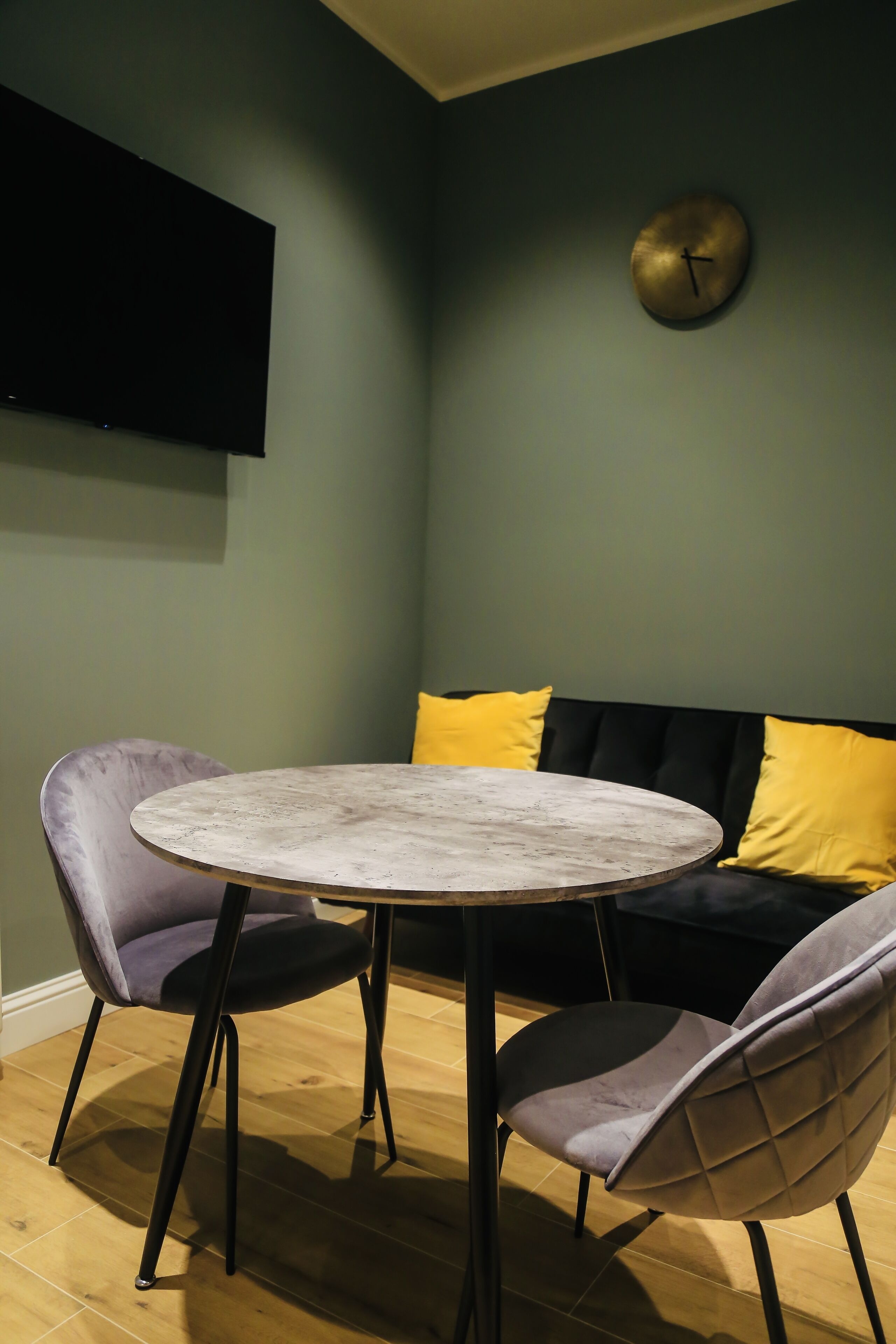 A room with a round table, two chairs, a sofa with yellow cushions, and a wall-mounted clock.