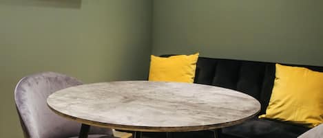 A room with a round table, two chairs, a sofa with yellow cushions, and a wall-mounted clock.
