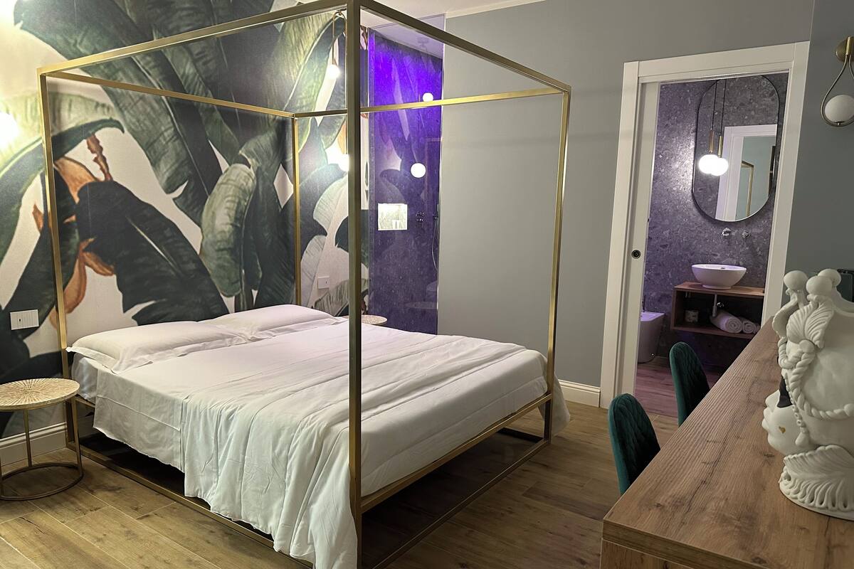 A four-poster bed with a white bedspread, a round wooden side table, a large abstract painting, a wooden dresser, and a bathroom with a sink and mirror.