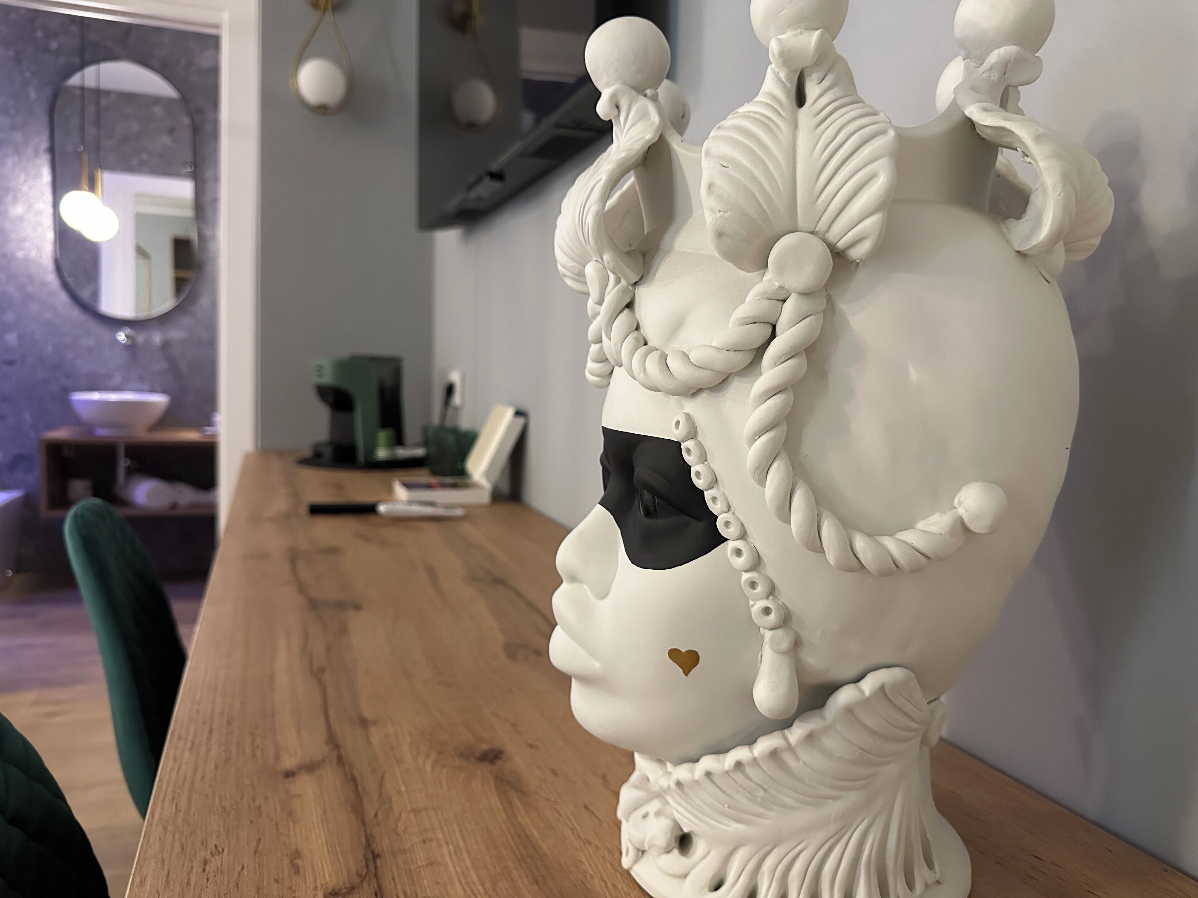 Un buste en céramique blanche, orné de motifs complexes, est posé sur une table en bois dans une pièce moderne, où l’on aperçoit un miroir, une machine à café et un fauteuil vert.